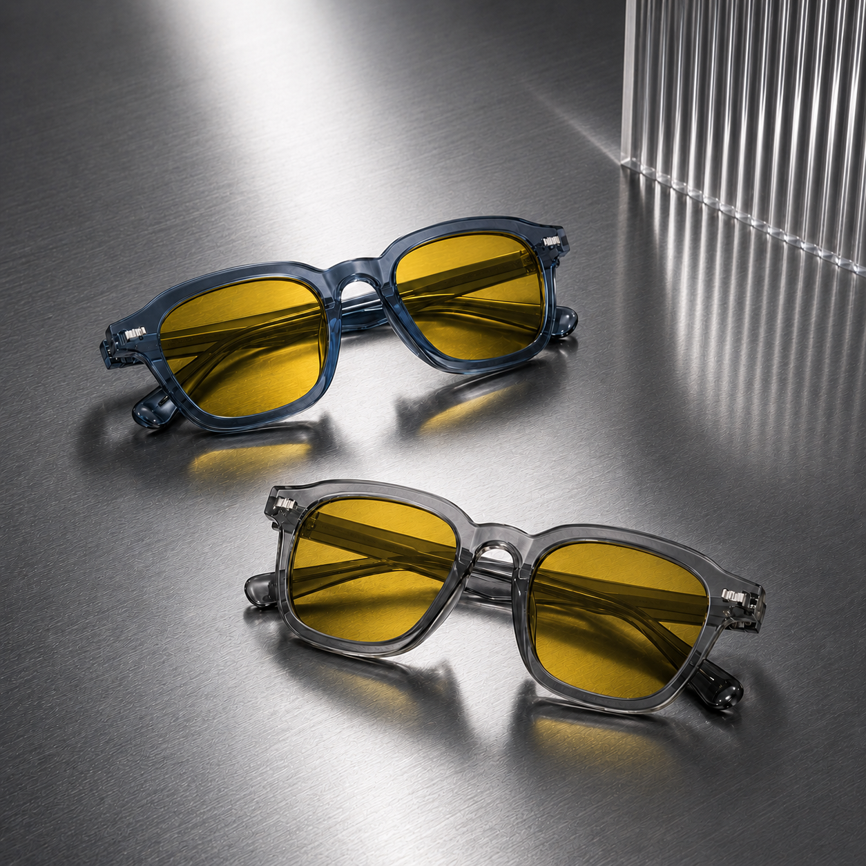 Óculos AURA Day Blue e Smoke com lentes amarelas com filtro de luz azul, dispostos sobre superfície metálica prateada.
