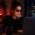 Mulher usando óculos AURA Night Blue com armação azul translúcida e lentes vermelhas enquanto trabalha no notebook à noite.