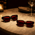 Óculos VESPER Night Black e Tortoise com lentes vermelhas, fechados sobre uma mesa de restaurante em ambiente noturno com iluminação aconchegante.