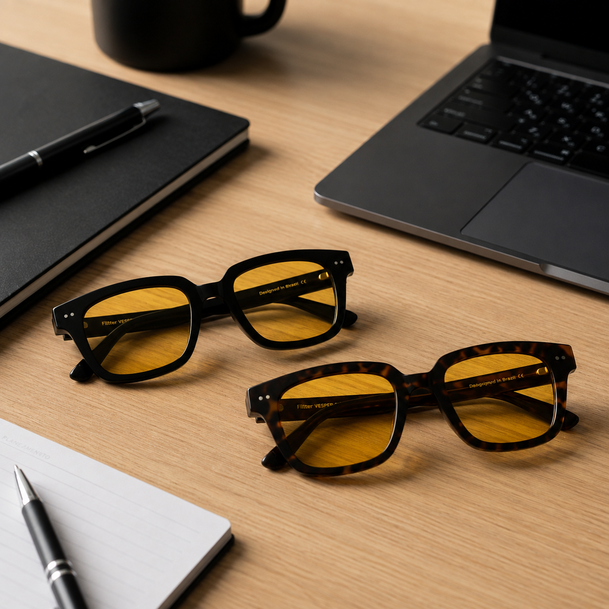 Óculos Vesper Day nas cores Black e Tortoise com lentes amarelas de filtro de luz azul, sobre mesa de escritório com notebook e acessórios de trabalho.